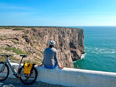 Cyclist with helmet sitting on white wall overlooking dramatic Algarve cliffs and turquoise Atlantic Ocean. Bicycle with yellow panniers nearby.
