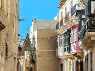 Smal gade i Rabat, Malta med traditionelle kalkstensbyggninger og farverige lukkede balkoner under klar blå himmel.