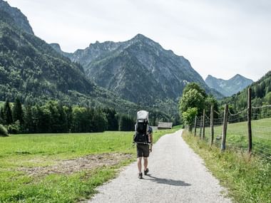 Vandrer med rygsæk på asfalteret sti gennem grøn dal i Salzkammergut, Østrig. Skovklædte bjerge rejser sig i baggrunden.