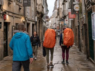 Pilgrimme med orange regnovertræk på rygsække går gennem smal stengade på Camino de Santiago. Historiske bygninger langs begge sider.