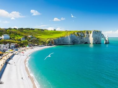 Flygvy över Étretats berömda vita kritklippor och naturliga klippbåge längs den turkosa kusten med stenstranden och kuststad.