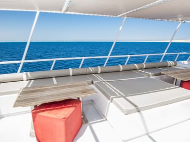 Hvidt skibsdæk på MV Tala med polstrede siddeområder, røde opbevaringskasser og panoramaudsigt over havet gennem beskyttelsesrækværk.