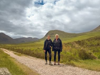 To vandrere på en grusvej på West Highland Way med grønne bakker og dramatisk overskyet himmel i de skotske højlande.