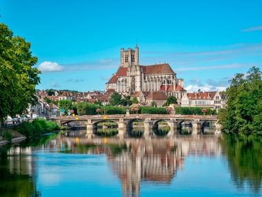 Den gotiske katedral i Auxerre dominerer horisonten over en stenbro, der krydser Yonne, med perfekte refleksioner i det stille vand.