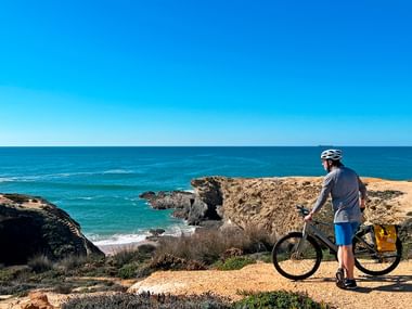 Syklist med hjelm står ved sykkel på gyllen klippe med utsikt over turkis Atlanterhav og steinete kyst i Portugal.