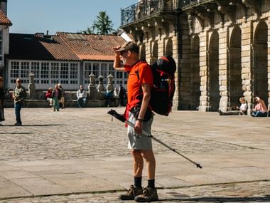 Pilgrim med rygsæk og vandrestav på brostenspladsen i Santiago de Compostela, med historisk katedral og traditionelle bygninger.