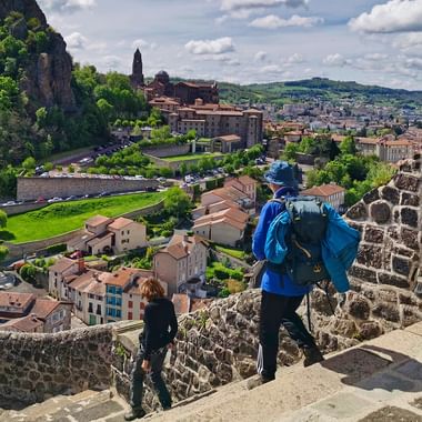 To vandrere med rygsække på stenmure ser ud over Le Puy-en-Velay. Den historiske by breder sig ud med røde tegltage.