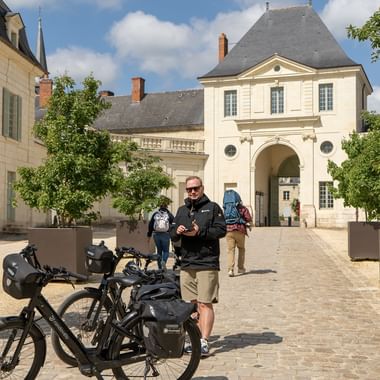 Cyklist med turistcykel på kullerstensborggård vid Loire-slott. Klassisk fransk arkitektur med välvd ingång synlig.