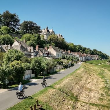 To cyklister på asfalteret sti langs Loire med historisk slot på bakketop over franske landsbyhuse under klar blå himmel.