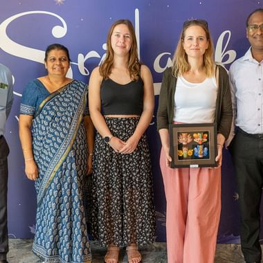 Fem personer poserer sammen indendørs mod en lilla væg med Sri Lanka branding. Gruppen inkluderer mænd i forretningsdragt og kvinder i afslappet påklædning.