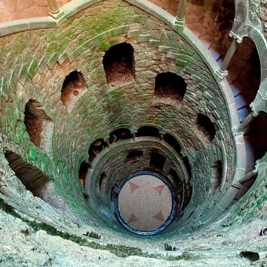 Flyfoto av en rund steinbrønn med spiraltrapp som fører nedover. Den gamle strukturen viser forvitrede steinvegger og buede åpninger.