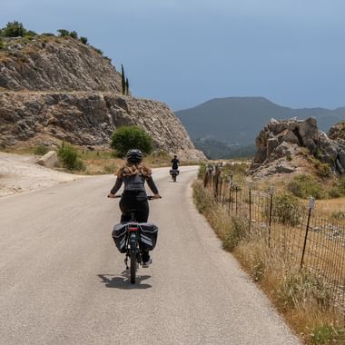 To cyklister med cykeltasker kører på en asfalteret bjergvej på Kefalonia, omgivet af klippevægge og middelhavs landskab.