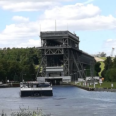 White passenger vessel sailing through raised steel lift bridge on calm waterway, surrounded by green trees and cloudy sky.