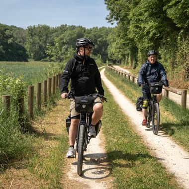 Två cyklister med hjälmar cyklar på en grusväg genom Loire-landskapet, omgivna av gröna fält och trästaket.