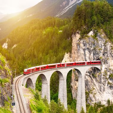 Rødt passagertog krydser en buet stenviadukt med flere buer gennem en schweizisk bjergdal med tætte skove og klippevægge.