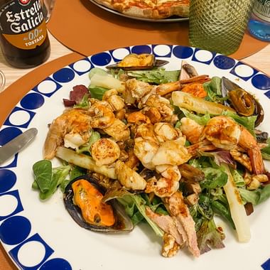 Plate with mixed seafood salad including shrimp, mussels, and white asparagus on greens. Beer bottle and glasses visible on wooden table.