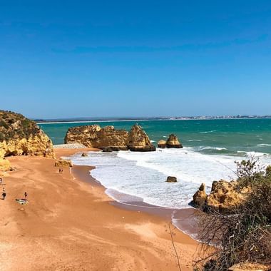 Scenic Algarve beach with golden sandstone cliffs, turquoise ocean, and rock formations. Visitors enjoy the sandy shore under clear blue sky.