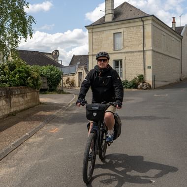 Manlig cyklist med hjälm och mörka kläder cyklar genom lugn byväg med traditionella franska kalkstensbyggnader.