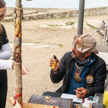 Kvinnelig pilegrim står ved siden av sittende mann som stempler pilegrimspass ved rasteplass på Portugisiske Camino. Kyststien og steinmurer synlige.