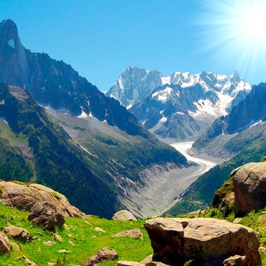 To vandrere står på klippefyldt terræn med udsigt over Mont Blanc massivet med snedækkede toppe, gletsjere og grønne alpenge.