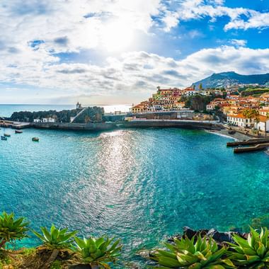 Panoramautsikt over en beskyttet bukt på Madeira med turkist vann, små båter, fargerike hus på åssider og tropisk vegetasjon.