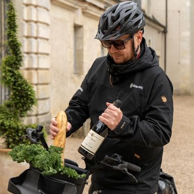 Cyklist i sort jakke og hjelm holder vinflaske og stokbrød, står med cykel foran elegant fransk slotsgård.