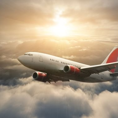 White commercial aircraft with red tail flying through dramatic cloudy sky with golden sunlight breaking through the clouds above.
