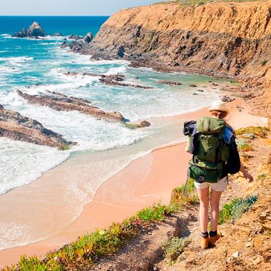 Fotturist med ryggsekk på klippesti med utsikt over turkis hav og sandstrand. Steinete kyst med dramatiske klipper i Alentejo.