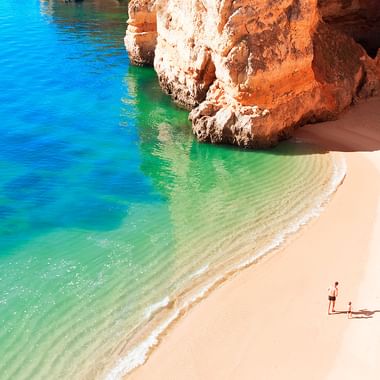 Aerial view of a secluded beach in Algarve with white sand, turquoise water, and dramatic orange limestone cliffs. Two people relax on the shore.