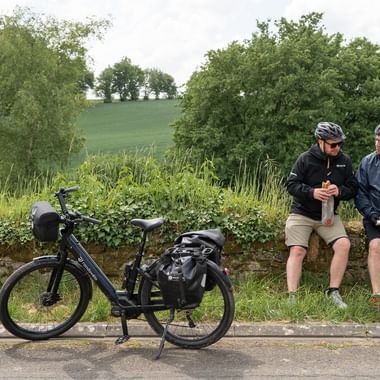 Två cyklister med hjälmar sitter på stenmur och äter mellanmål, med turistcyklar parkerade i grönt landskap.