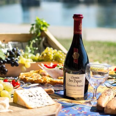 Udendørs picnic med vinflasker, ost, druer, brød og trækasse på ternet tæppe i solrig landskab.