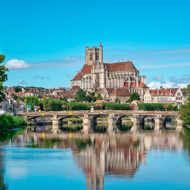 Den gotiska katedralen i Auxerre dominerar horisonten ovanför en stenbro över Yonne, med perfekta reflektioner i det lugna vattnet.
