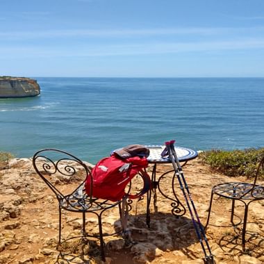 Black wrought iron table and chairs on clifftop overlooking turquoise ocean and golden cliffs in Algarve. Red backpack sits on table.