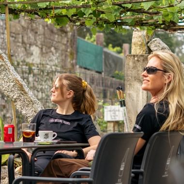 To kvinner sitter på en utendørs kafé under en vinbevokst pergola på den Portugisiske Camino. Kaffekopper og drikker på bordet.