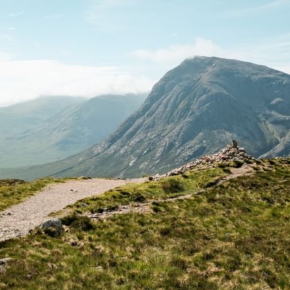 Grusvandrerute på West Highland Way med grønne græsskråninger til dramatiske bjergtinder under blå himmel i Skotland.