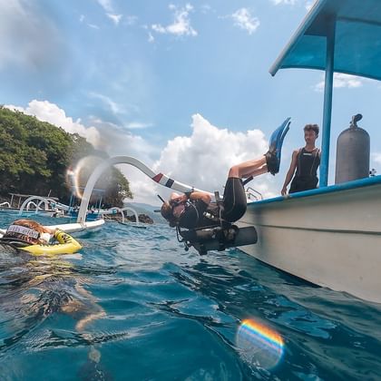 Dykker i våtdrakt går i turkist vann fra hvit dykkerbåt på Bali. Dykkerflasker synlige på båten, grønn kystlinje og blå himmel i bakgrunnen.