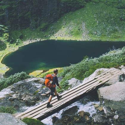 Vandrer med orange rygsæk krydser træbro over bjergbæk i Aurlandsdalen, med mørk bjergsø og grønne skråninger nedenfor.