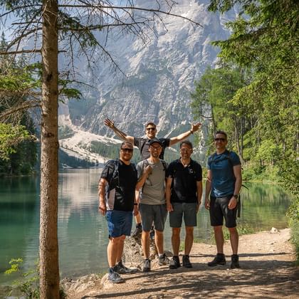 Fem vandrere poserer ved en turkis bjergsø i Dolomitterne, omgivet af tæt skov og dramatiske klippetoppe.