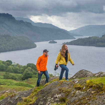 Fotturister på West Highland Way ved innsjø To fotturister i fargerike jakker står på mosegrodd stein med utsikt over en skotsk innsjø omgitt av grønne åser og fjell.