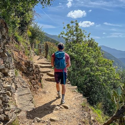 Hiker with blue backpack walking on stone path along Ligurian coast. Stone wall on left, Mediterranean Sea and mountains visible on right.