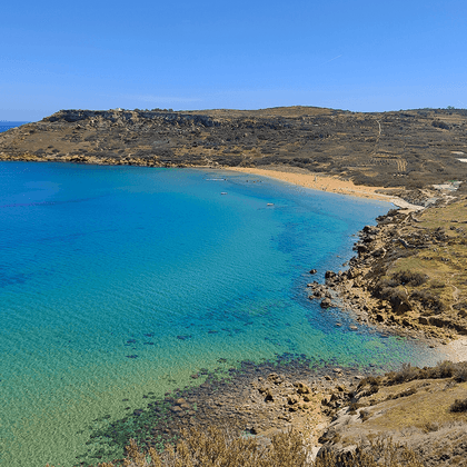 Luftfoto af Ramla Beach på Gozo med turkisblåt vand, gylden sandstrand og klippefyldte skråninger under klar himmel.