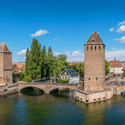 Middelalderlige stentårne flankerer en bro over en kanal i Strasbourg. Historiske bygninger og kirketårne synlige i baggrunden under blå himmel.