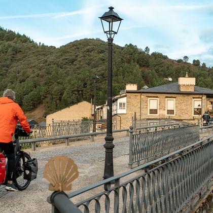 Cyklist i orange jakke med lastet turistcykel krydser stenbro med udsmykkede rækværk. Camino-muslingesymbol synligt på brogelænder.