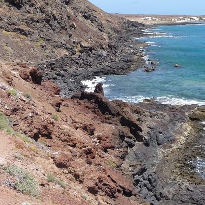 Hiker walking on volcanic rocky coastline of La Graciosa with turquoise ocean waters and distant white buildings visible on the horizon.