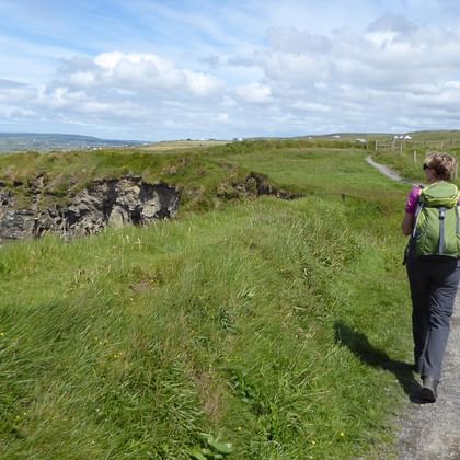 Vandrer med rygsæk går på grusvej langs stenmur i irsk landskab. Grønne marker, fjerne bakker og skyet himmel synlig.