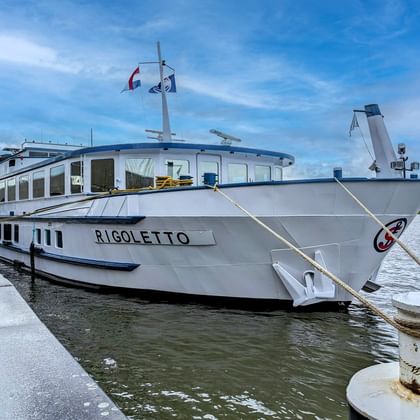 Hvidt flodkrydstogtskib MS Rigoletto fortøjet ved betonkaj med fransk flag på mast, flere dæk med vinduer, under blå himmel.