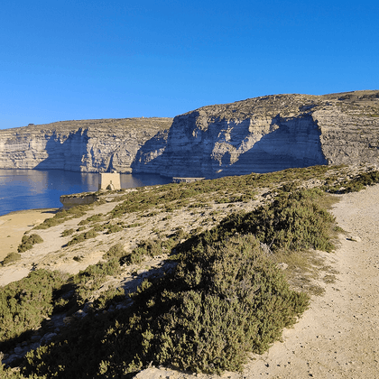 Grusvej langs Middelhavets kyst på Gozo med dramatiske kalkstensklipper, sandstrand og blåt hav under klar himmel.