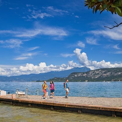 Lake Garda wooden pier with hikers Three hikers on wooden pier at Lake Garda with flagpole, mountains in background under blue sky with white clouds.
