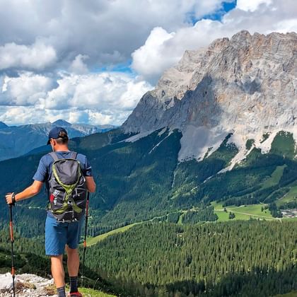 Vandrer med rygsæk og vandrestave ser på dramatiske Zugspitze toppe, grønne dale og skove under overskyet himmel.