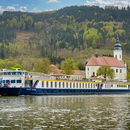 MS Prinzessin Katharina flodkrydstogtskib sejler på stille vand med hvid kirke og skovklædte bakker i baggrunden under blå himmel.